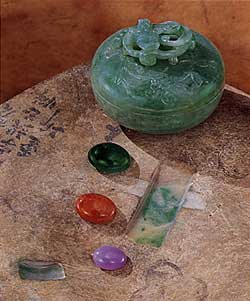 Figure 1. "Windows" cut into this otherwise undistinguished boulder from the Burma Jade Tract reveal the presence of a rich green in the jadeite beneath the skin. Boulders such as this are the source of the fine green, orange-red, and lavender cabochons that are much sought-after in China and elsewhere. The bowl is approximately 6 cm wide x 5 cm high. The cabochons measure approximately 15 x 19 mm (green), 13 x 18 mm (orange-red), and 10 x 14 mm (lavender). Courtesy of Bill Larson and Pala International; photo © Harold & Erica Van Pelt.