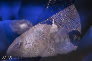 In this sapphire from Sri Lanka, evidence of high temperature heat treatment can be found in this moiré-patterned fingerprint. The once-lovely lacy pattern of liquid droplets is now besmirched by circular “explosions,” where the pressure from heating caused ruptures in the microscopic negative crystals, thus deflowering Mother Nature’s exquisite work of art.