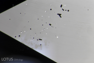 This 27+ carat sapphire came in for testing. Microscopic examination quickly revealed that it was titanium diffused, with unusual patches of blue color bleeding from inclusions. Of course a natural 27-ct sapphire of good clarity would never be used for such treatment, because the natural untreated sapphire would be worth far more. The reality was that synthetic sapphire was used for the treatment. This image shows surface pits, along with unusual metallic inclusions cut through on the surface.